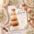 A styled flat-lay of a bridal shower invitation card featuring a three-tier coffee cake illustration and the text 'Love is Brewing.' The card is surrounded by cream roses, baby's breath, coffee beans, a satin ribbon, and a pearl-covered dish on a neutral fabric background.